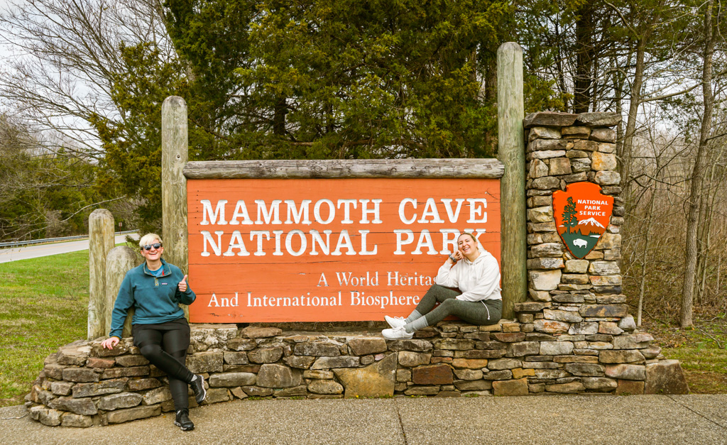 orange sign reads Mammoth Cave National Park A World Heritage Site And International Biosphere. Alex sits to the left giving a thumbs up and Chris sits the right.