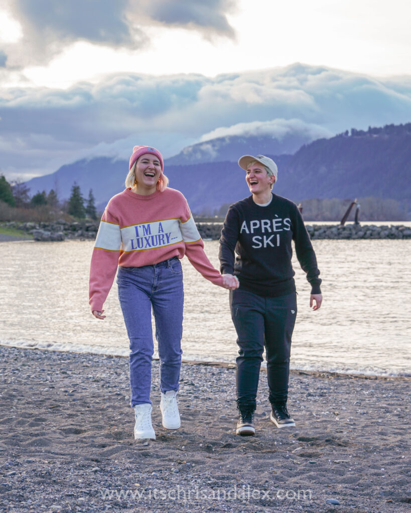 Sapphic couple Chris and Alex walk holding hands at the waterfront of the Columbia River in Hood River, Oregon with Washington behind them.